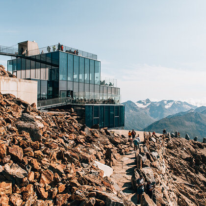 A modern glass building towers on a rocky mountain ridge with people moving across the scene. Snow-capped peaks are in the background under a clear sky.