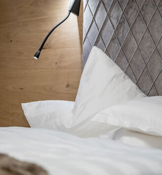 A neatly made bed with white bedding, a padded gray headboard with a diamond pattern and a modern black wall-mounted reading lamp against a wooden wall.