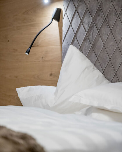 A neatly made bed with white bedding, a padded gray headboard with a diamond pattern and a modern black wall-mounted reading lamp against a wooden wall.
