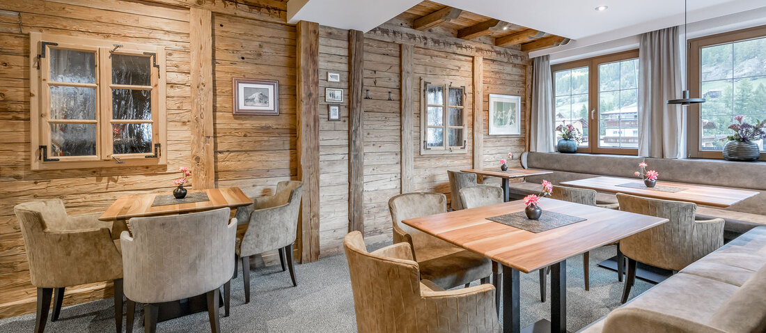 Cozy bar interior with wood-paneled walls, light brown upholstered armchairs, wooden tables, gray carpeting, large windows and small vases of flowers on the tables. The room is flooded with natural light. 