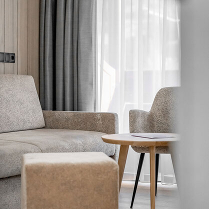 Modern living room with a beige sofa, a light-wood side table and an upholstered armchair near large windows with sheer curtains and dark drapes.
