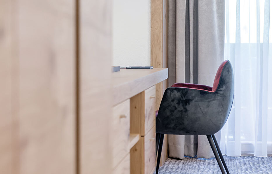 Modern interior featuring a desk, a black chair with a red seat cushion, a pendant light, beige curtains and natural light streaming in through a clear window. Neutral tones and minimalist design.
