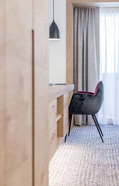 Modernes Interieur mit einem Schreibtisch, einem schwarzen Stuhl mit rotem Sitzkissen, einer Hängeleuchte, beigen Vorhängen und natürlichem Licht, das durch ein transparentes Fenster hereinfällt. Neutrale Farbtöne und minimalistisches Design.
