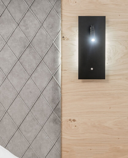 An einer hellen Holzwand neben einem grauen, mit Rautenmuster bezogenen Kopfteil ist eine moderne Wandleuchte mit einer kleinen Lampe angebracht.