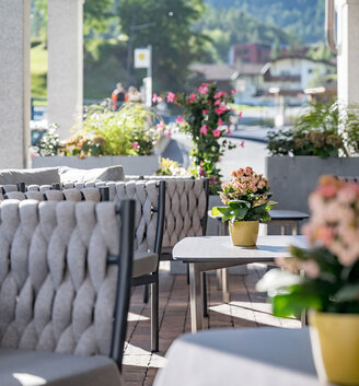 Outdoor terrace featuring grey cushioned chairs and tables adorned with potted flowers. Bright sunlight, plants and mountains are in the background. The setting is cozy and inviting.