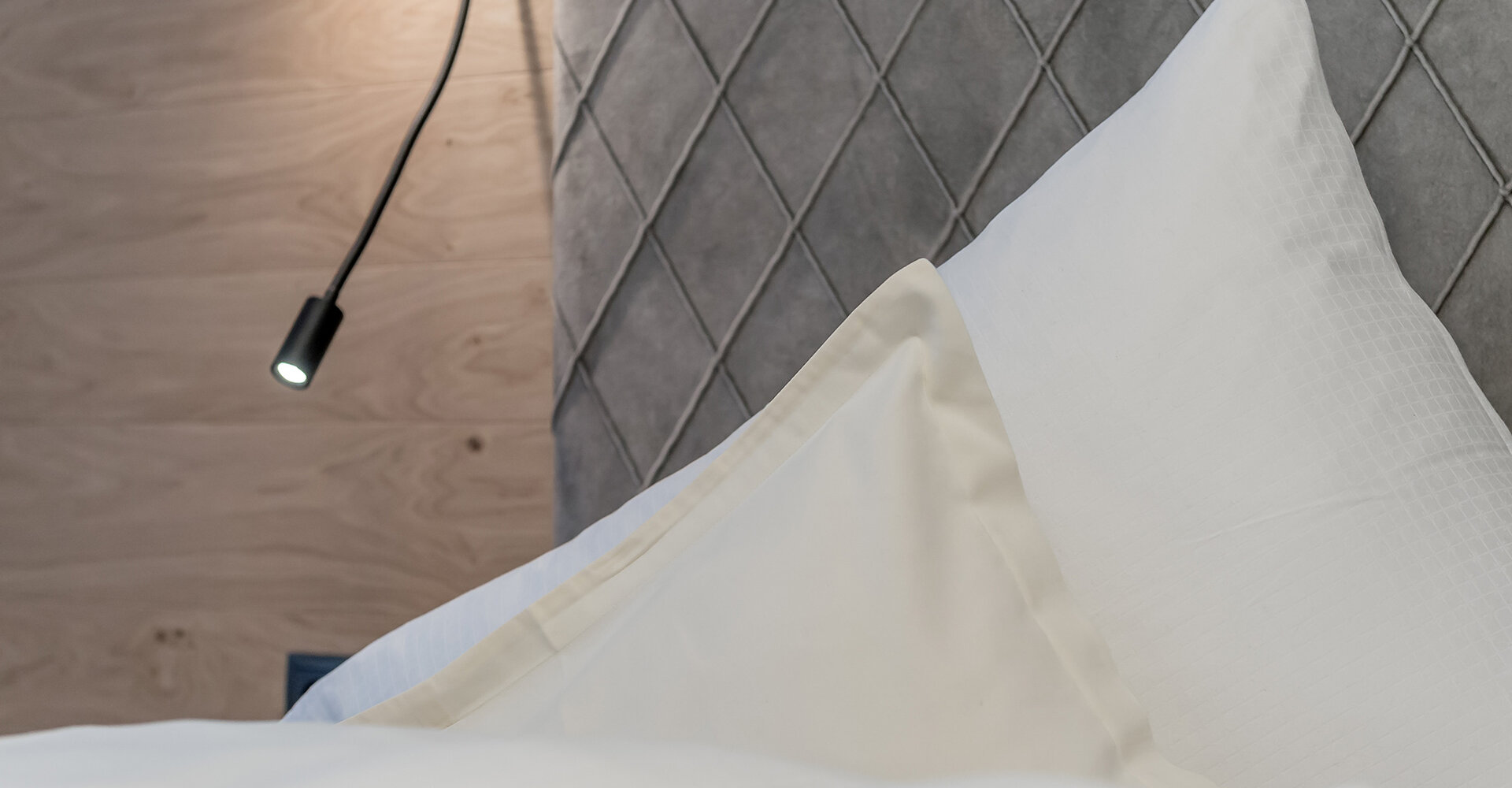 Eine Nahaufnahme eines ordentlich gemachten Bettes mit weißen Kissen und einem gesteppten grauen Kopfteil. An einer holzvertäfelten Wand neben dem Bett ist eine schwenkbare Leselampe angebracht.