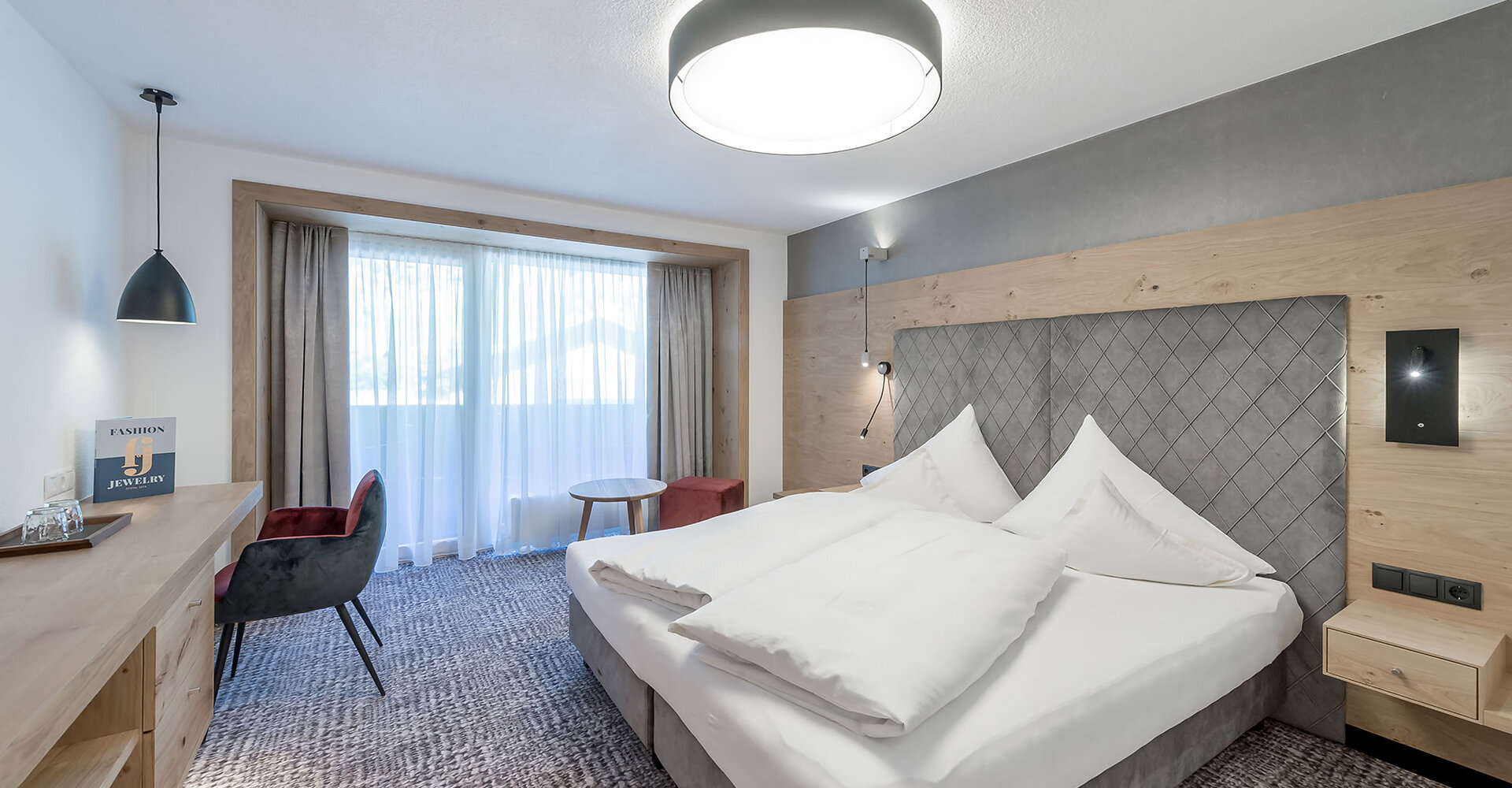Ein modernes Hotelzimmer mit einem großen Bett, hellen Holzmöbeln, einem kleinen runden Tisch mit zwei Stühlen und einem großen Fenster mit transparenten Vorhängen, durch die Tageslicht hereinströmt.