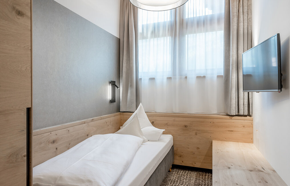 A small, modern bedroom with a single bed, light wood furnishings and grey tones, a wall lamp, a wall-mounted flat-screen TV and a large window with sheer curtains allowing daylight to stream in.