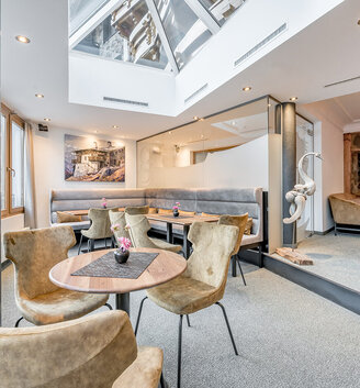 Modern lounge with round tables and beige chairs, large windows and a skylight. There is a bench along the back wall. The room is bright and inviting, featuring neutral colors and modern furnishings.