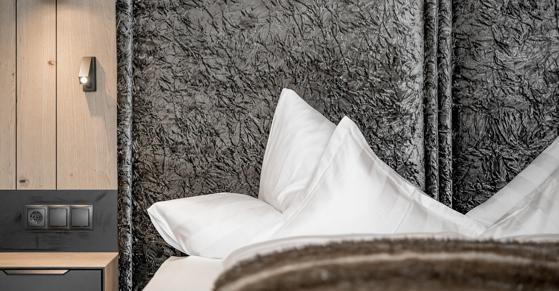 A close-up of a modern hotel bed with white pillows, a grey textured headboard, a faux fur blanket, a wooden bedside table with electrical sockets and a black wall lamp.