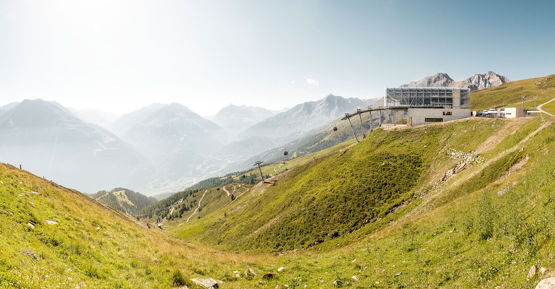 Ein Panoramablick auf grüne Hügel mit einer modernen Seilbahnstation am Hang, Seilbahnen, die den Berg hinauffahren, und dunstige, weit entfernte Berge unter einem klaren Himmel.
