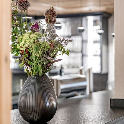 On a dark counter in a modern bar stands a striped vase with colorful flowers. In the background, there are shelves with bottles and glasses as well as a coffee machine.