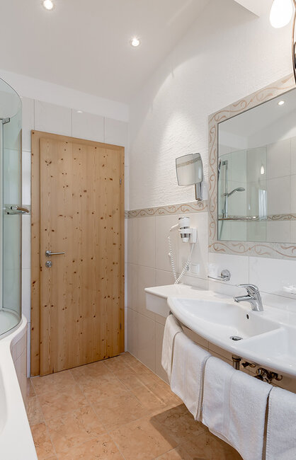 Modernes Badezimmer mit Badewanne, Glasduschkabine, Doppelwaschbecken, großem Spiegel, weißen Fliesen, einer Tür aus hellem Holz und Handtüchern, die unter den Waschbecken hängen.