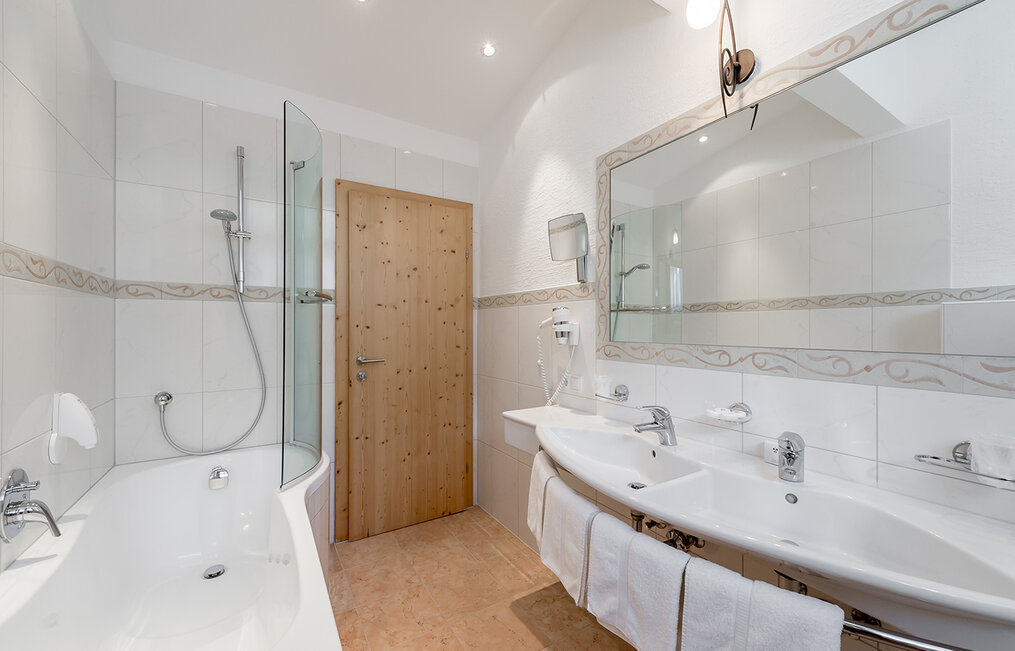 Modern bathroom with a bathtub, glass shower enclosure, double washbasin, large mirror, white tiles, a light-wood door and towels hanging beneath the sinks.
