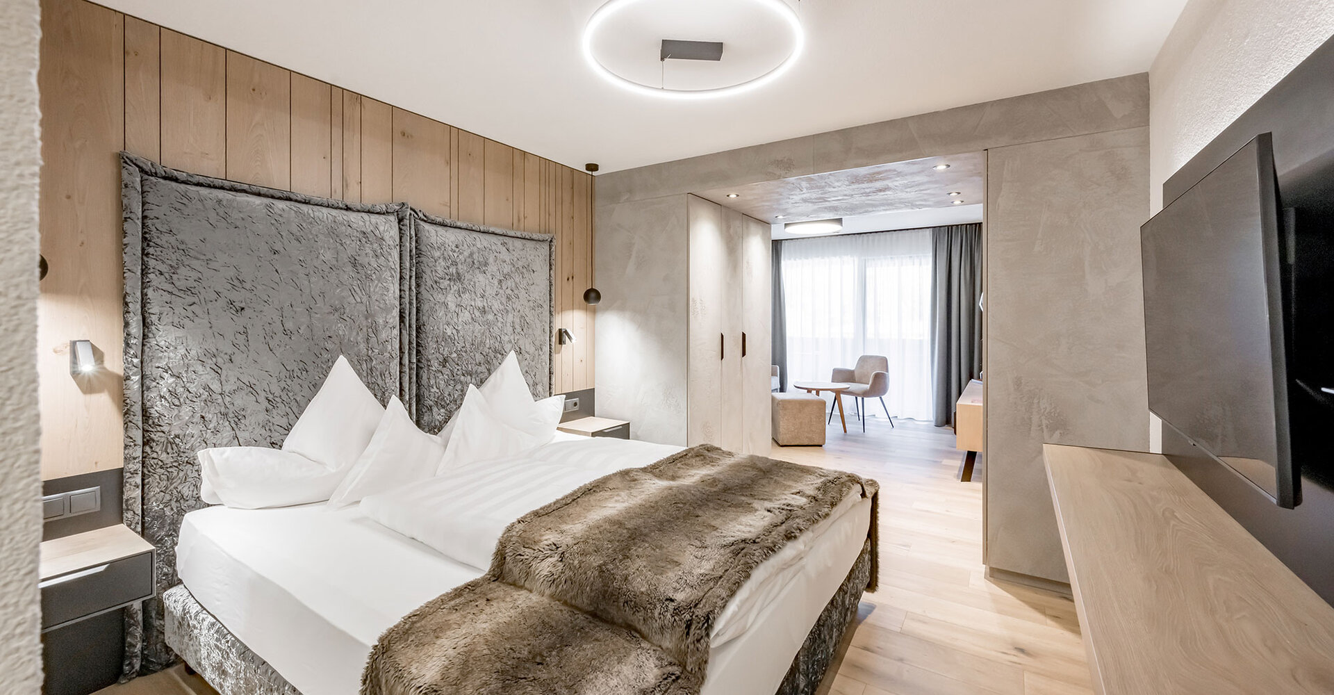 Modern hotel room featuring a large bed, wooden elements, a TV and a workspace next to a bright window. Neutral tones and soft lighting create a cozy, inviting atmosphere.