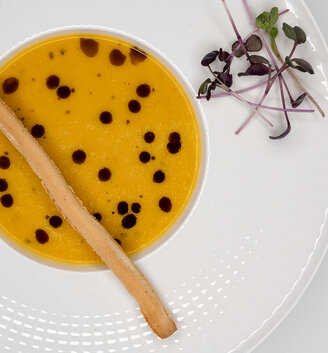 A white plate containing yellow soup with dark droplets floating on the surface, garnished with a long breadstick and microgreens.