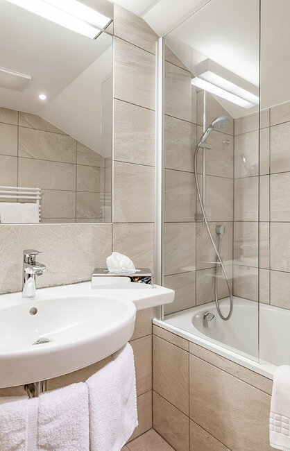 Modernes Badezimmer mit hellbeigen Fliesen, einem weißen Waschbecken, einem großen Spiegel, einem an der Wand montierten Handtuchwärmer und einer Badewanne mit einer Duschabtrennung aus Glas. Handtücher hängen am Waschbecken und an der Badewanne.
