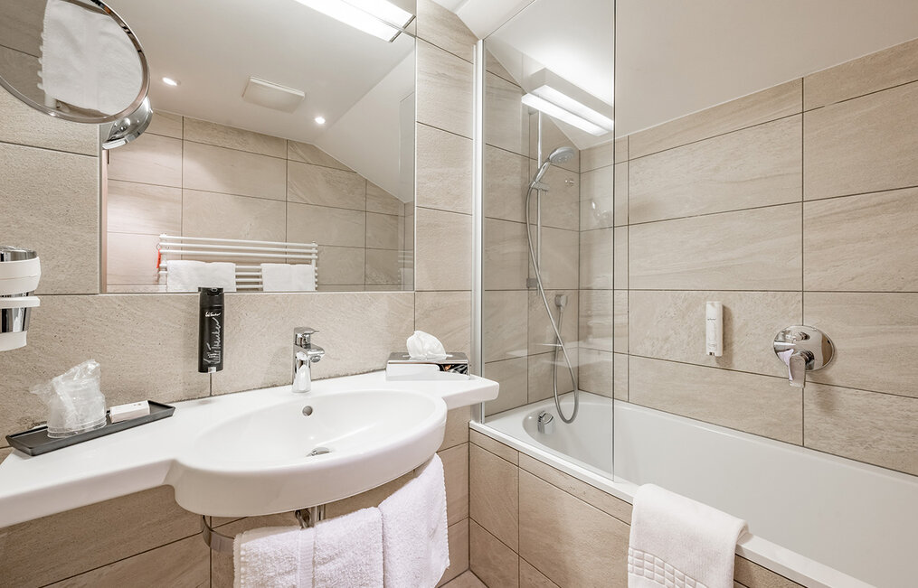 Modern bathroom with light beige tiles, a white sink, a large mirror, a wall-mounted towel warmer and a bathtub with a glass shower screen. Towels are hanging next to the washbasin and the bathtub.