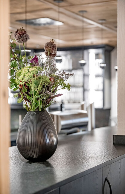 Eine dunkle, gerippte Vase mit verschiedenen Wildblumen steht auf einer grauen Arbeitsplatte in einer modernen Küche mit Holzakzenten und hängenden Pendelleuchten im Hintergrund.