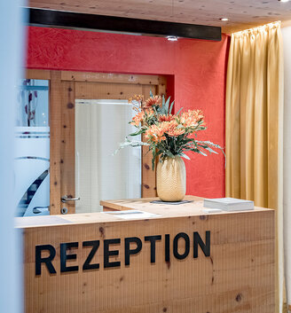 A wooden reception desk with the inscription "Rezeption," a vase with orange flowers, a wooden door, a red wall and yellow curtains in the background.
