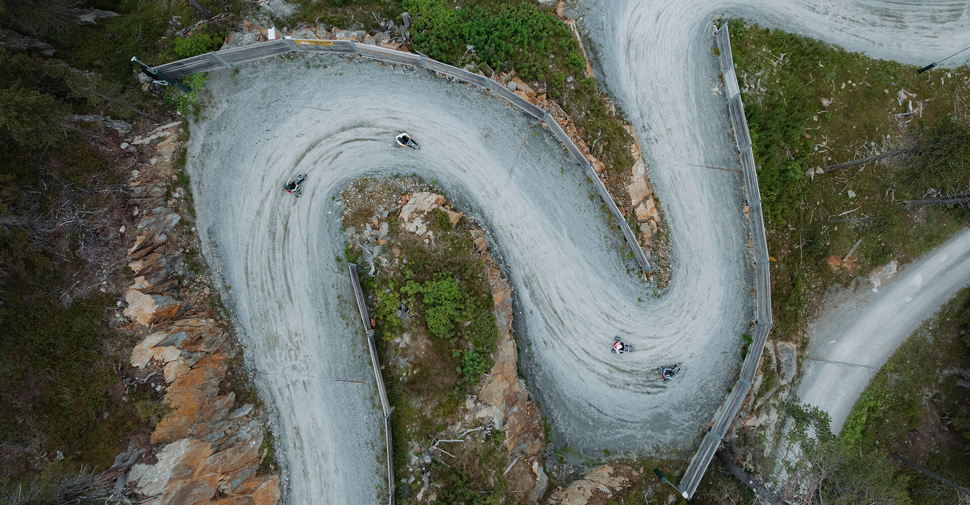 A winding road with sharp bends, lined with rocks and trees from a bird's-eye view. Several tricycle riders are navigating the hairpin turns.