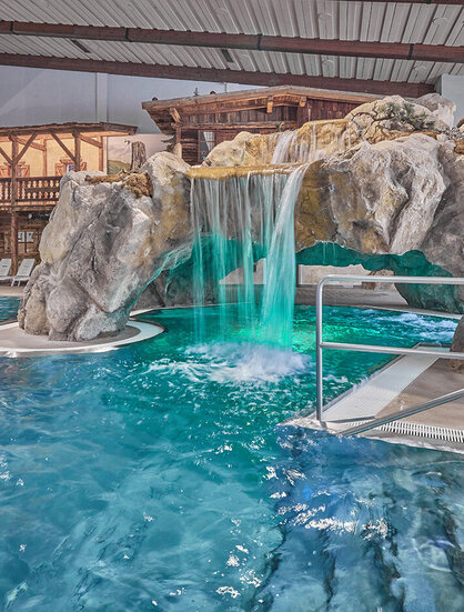 Hallenbad mit einer Felshöhlenkonstruktion, einem Wasserfall und einer Rampe, die ins Wasser führt. An einer Seite stehen Liegestühle.