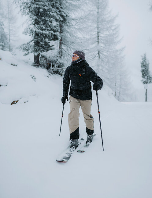 Eine Person in Winterkleidung und mit einer Mütze läuft mit Skiern bergauf durch eine verschneite Waldlandschaft. Schnee bedeckt den Boden und die Bäume und schafft so eine friedliche Winterkulisse.