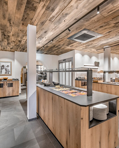 A modern buffet area featuring wooden cabinets and a wooden ceiling, grey tiled flooring and a varied breakfast selection arranged on the countertops. Shelving and tableware are visible, creating a neat and inviting dining atmosphere.