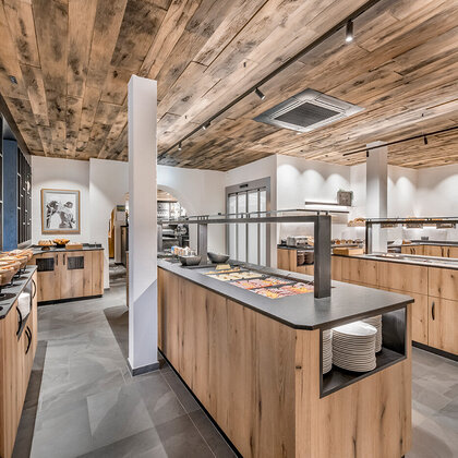 A modern buffet area featuring wooden cabinets and a wooden ceiling, grey tiled flooring and a varied breakfast selection arranged on the countertops. Shelving and tableware are visible, creating a neat and inviting dining atmosphere.