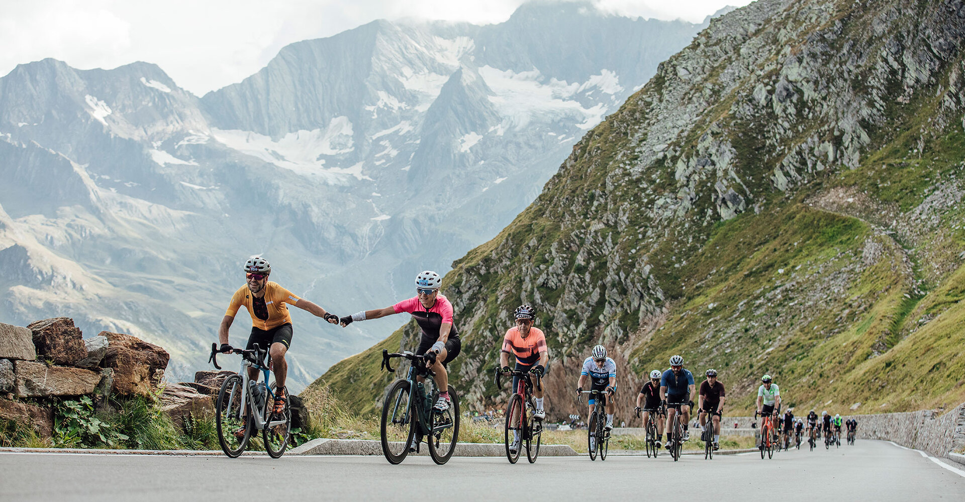 Cyclists ride up a mountain road, the two in front holding hands. Green hills and mountains line the winding road and even more cyclists follow them.