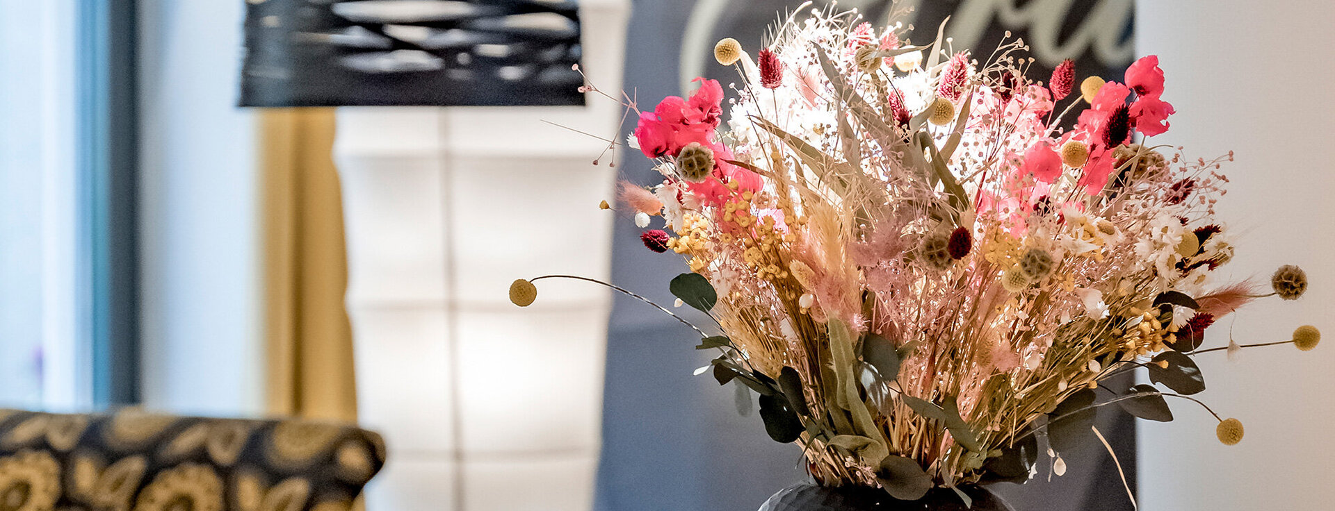 A bouquet of dried flowers with pink and yellow accents stands in a black vase. Behind it are a patterned chair, a black lamp and a wall with lettering.