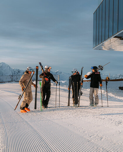 Four people in winter clothing are standing on groomed snow, holding skis and poles and chatting near a modern glass building. Snow-capped mountains in the background.
