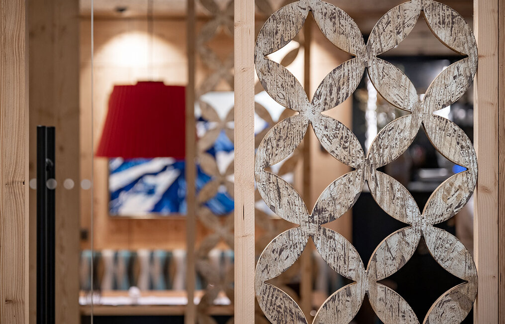 The focal point is a wooden partition element featuring a geometric floral pattern. Behind it, within a cozy, warmly lit interior, there are a red lampshade, an abstract artwork and a patterned bench.