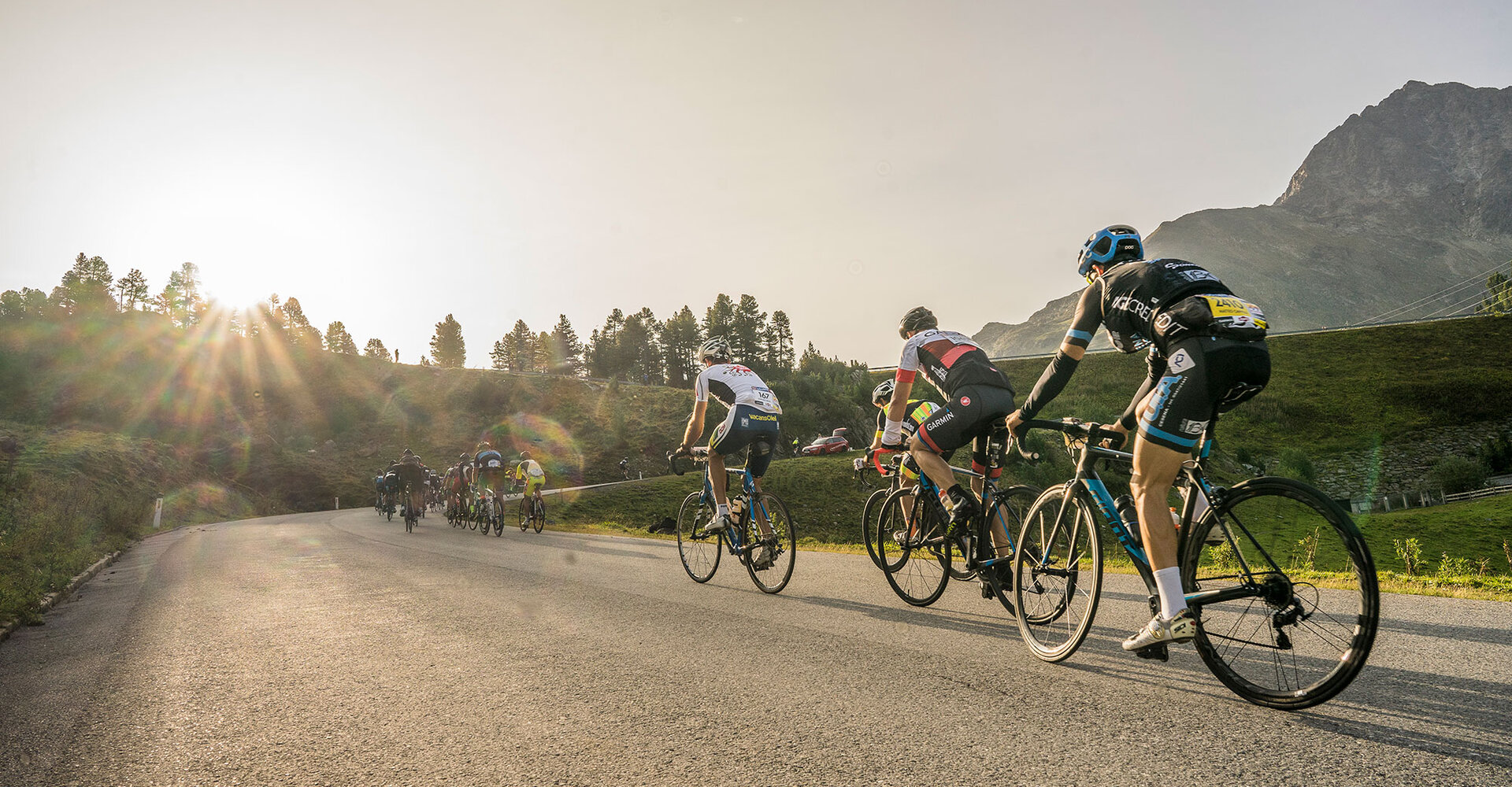 Cyclists ride uphill on an asphalt road at sunrise with mountains, trees and sunlight in the background. The cyclists are wearing helmets and cycling gear and are participating in a race.