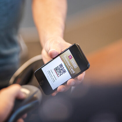 One person is holding a smartphone that displays a QR code labeled "Summer Card" on the screen, while another person scans it with a handheld device.