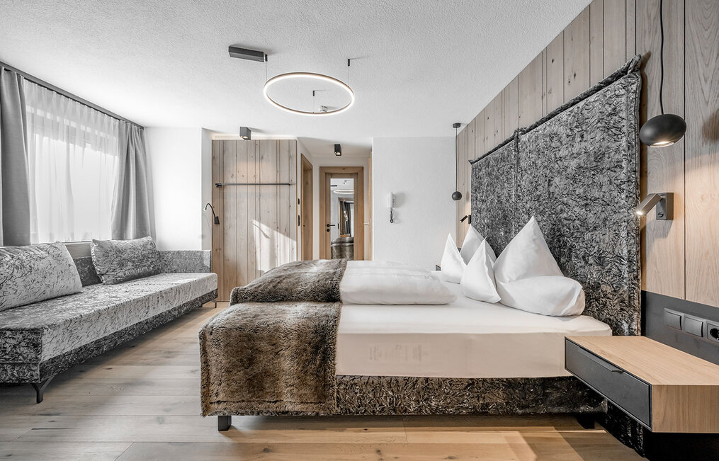 Modern hotel room featuring a large bed, a matching sofa, wooden floors and wood-paneled walls. Soft daylight streams in through a window with light-colored curtains. A minimalist, cozy and inviting design.