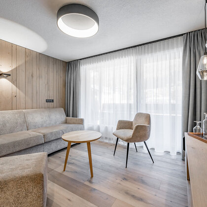 Modern living room with light wood floors, a beige sofa, an armchair, a round coffee table, a wooden accent wall, pendant lights and a large window with sheer curtains.