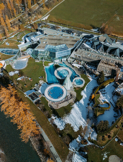 Aerial view of a modern resort with several outdoor pools, green lawns and glass buildings, surrounded by trees, patches of snow and a nearby river.