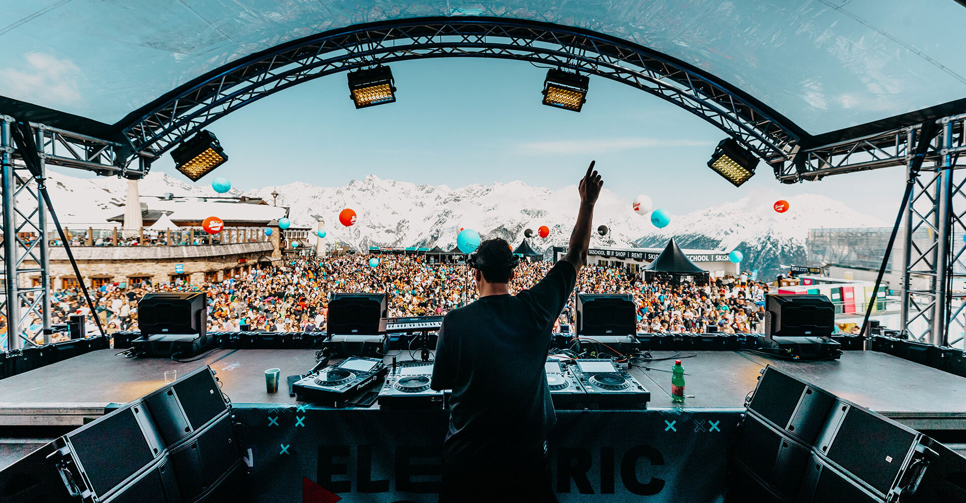 A DJ performs on stage at an open-air music festival in the mountains in front of a large crowd with colorful balloons and snow-covered mountain peaks in the background.