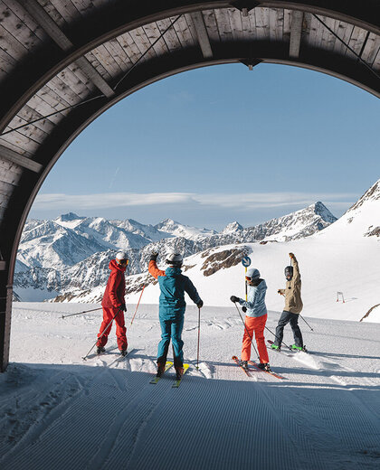 Four skiers stand in front of a wood-paneled tunnel in the snow-covered mountains, gazing out at the picturesque alpine landscape under a brilliant blue sky.