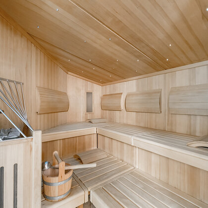 Moderne Holzsauna mit gestuften Bänken, einem Eimer mit einer Schöpfkelle und einem Metallofen in der Ecke. Der Raum wird durch kleine Deckenleuchten hell beleuchtet, was eine klare und entspannende Atmosphäre schafft.