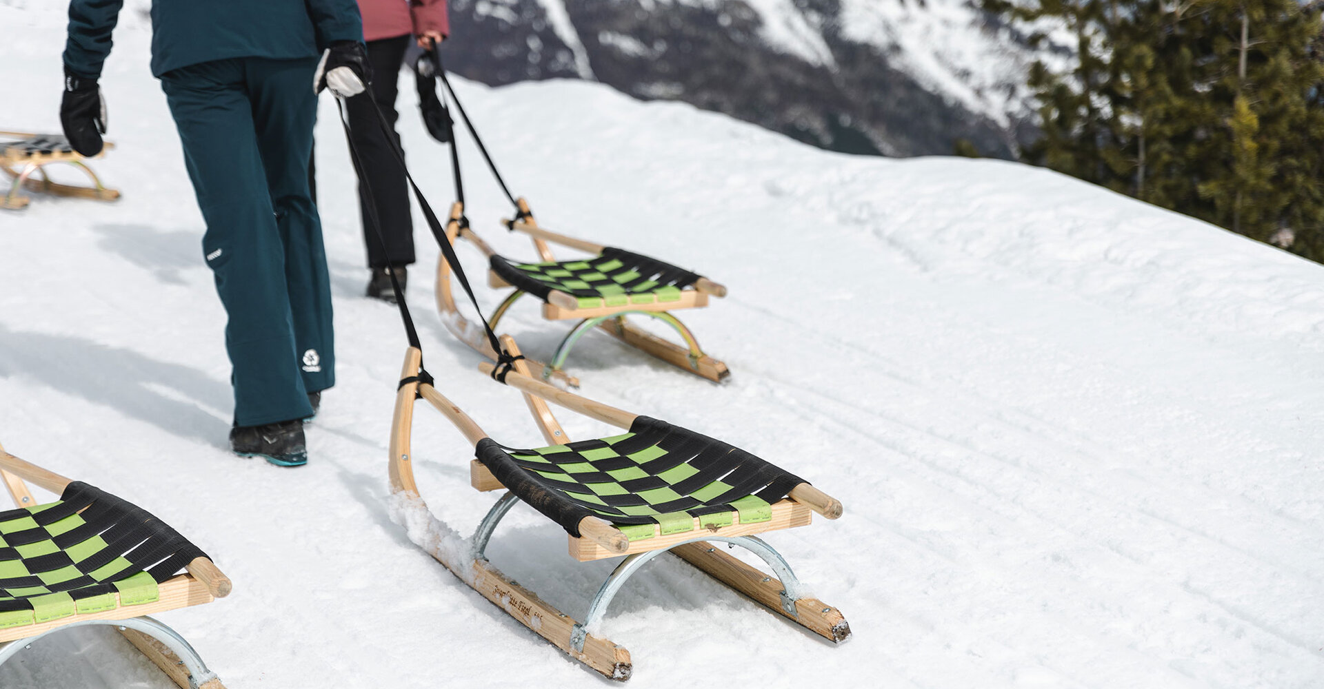 Two people are pulling wooden toboggans with green and black woven seats on a snowy mountain path. Snow-covered peaks and trees are in the background.
