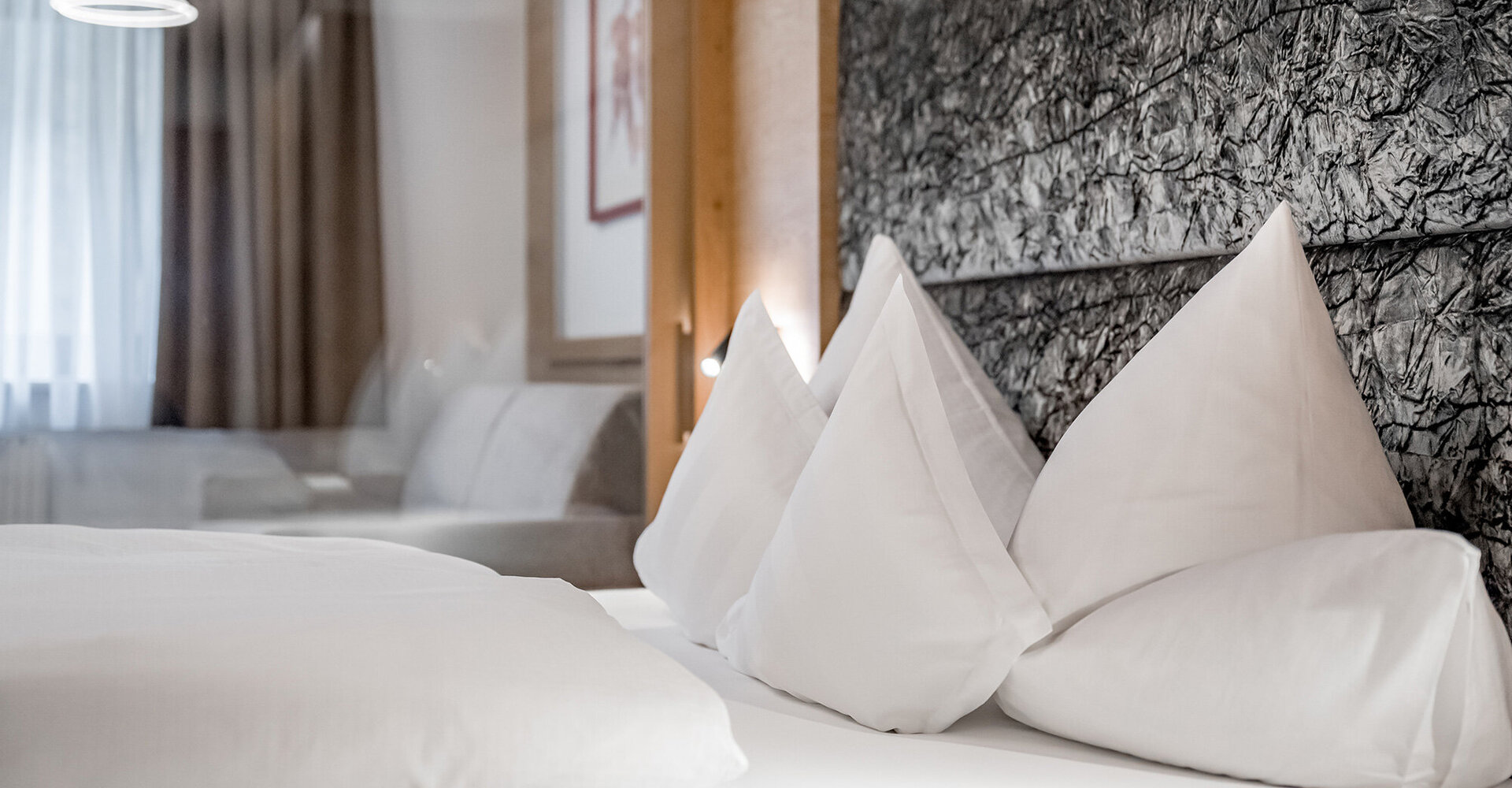 Ein ordentlich gemachtes Bett mit strahlend weißer Bettwäsche und vier dreieckigen Kissen, das vor einem strukturierten, dunklen Kopfteil in einem hellen, modernen Zimmer steht.