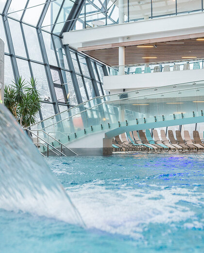 A large indoor pool with a winding waterfall, glass walls and ceiling, a bridge-like walkway, rows of free-standing loungers and streaming daylight.