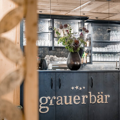 In the foreground, a stylish bar area featuring dark gray cabinets, a black vase with flowers, shelves filled with glasses and a wooden wall with a floral pattern.