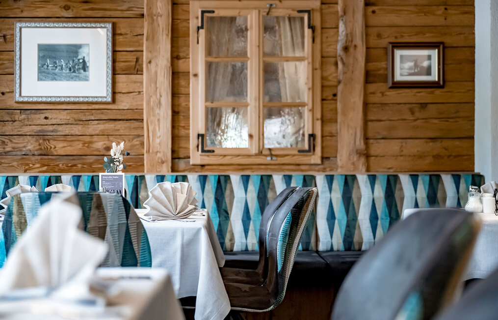 A cozy restaurant featuring wood-paneled walls, framed pictures and neatly set tables with white tablecloths and folded napkins. Blue-patterned upholstery adorns the seating area beneath a wooden window.
