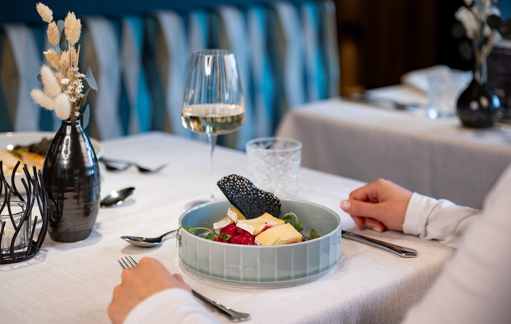 A person is sitting at a set dining table with a plate full of varied delicacies, a glass of white wine and a vase of dried flowers.