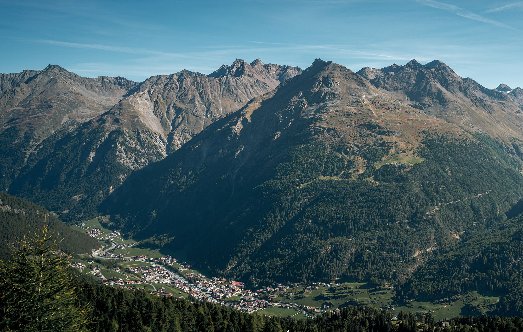 A small village lies in a green valley, surrounded by high, rugged mountains and beneath a brilliant blue sky.