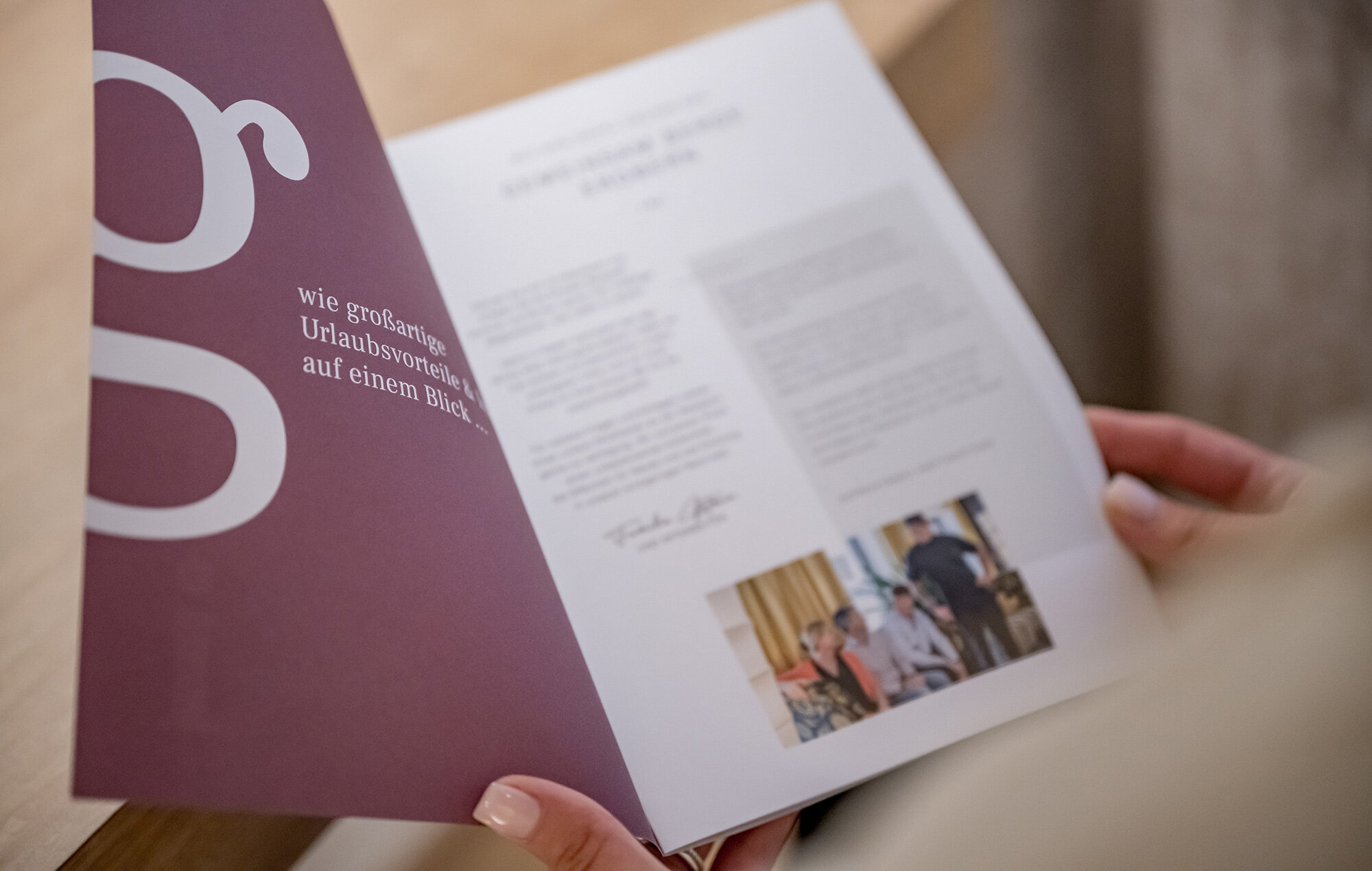 A person is holding a brochure and reading it. The brochure has a dark red cover and white lettering and displays a small photograph on the inside next to the printed text.