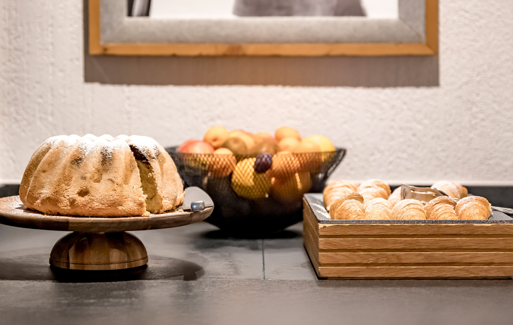 A ring cake with a slice cut out of it stands on a countertop next to a tray of pastries and a wire basket filled with apples and other fruit.